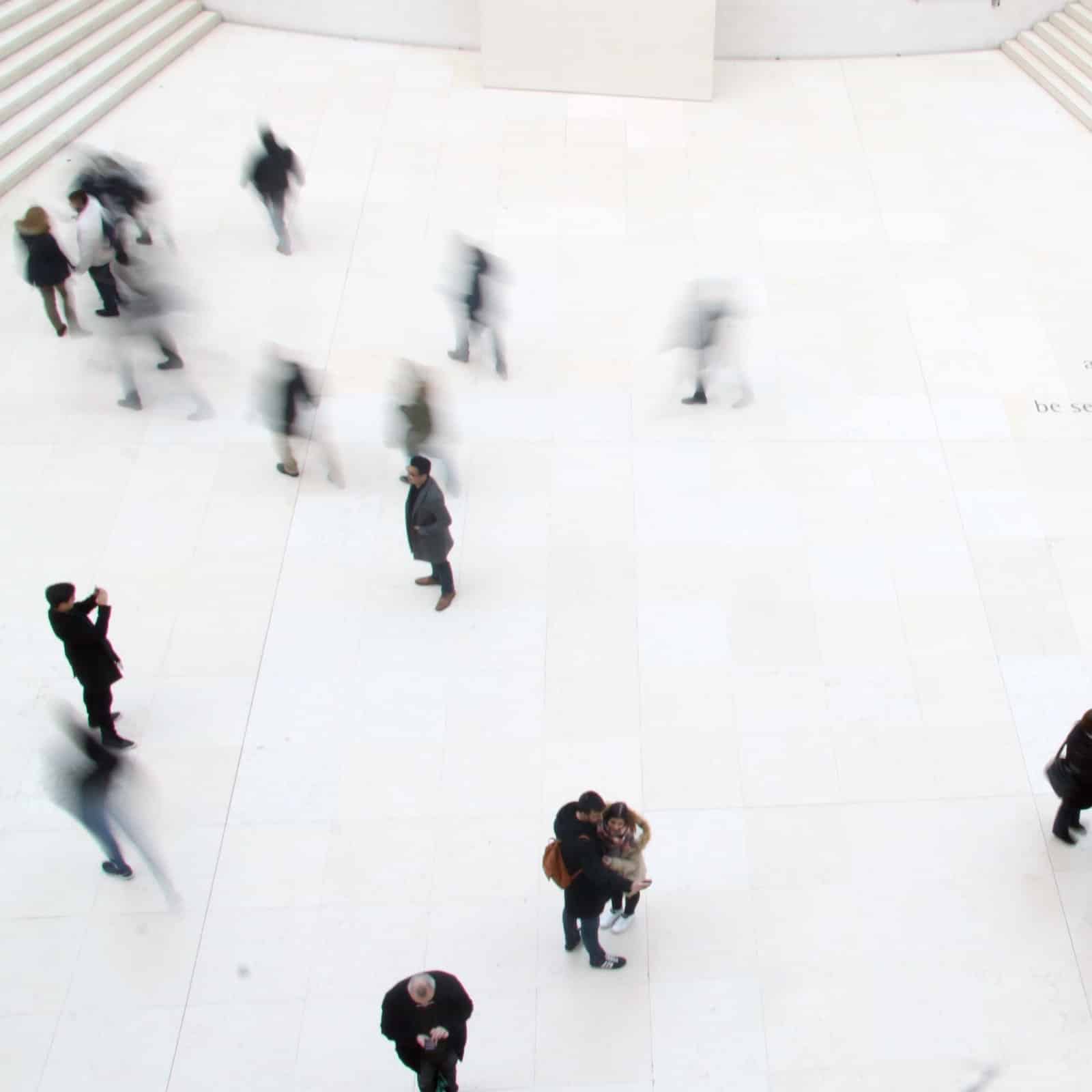 An image of a group of people moving through a building with white marble floors, view from above. The collective energy of the space is buzzing.