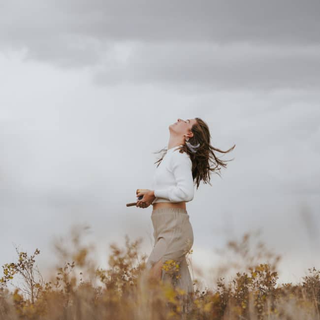 Erin Vivian standing in a field under a cloudy sky, holding a singing bowl. She feels lighter after a heart wall removal.