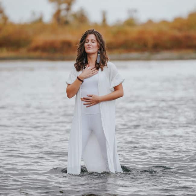 Erin Vivian is standing in the river with one hand on her heart and one on her belly. She is a certified emotion code and body code practitioner and created Waters Edge Healing.