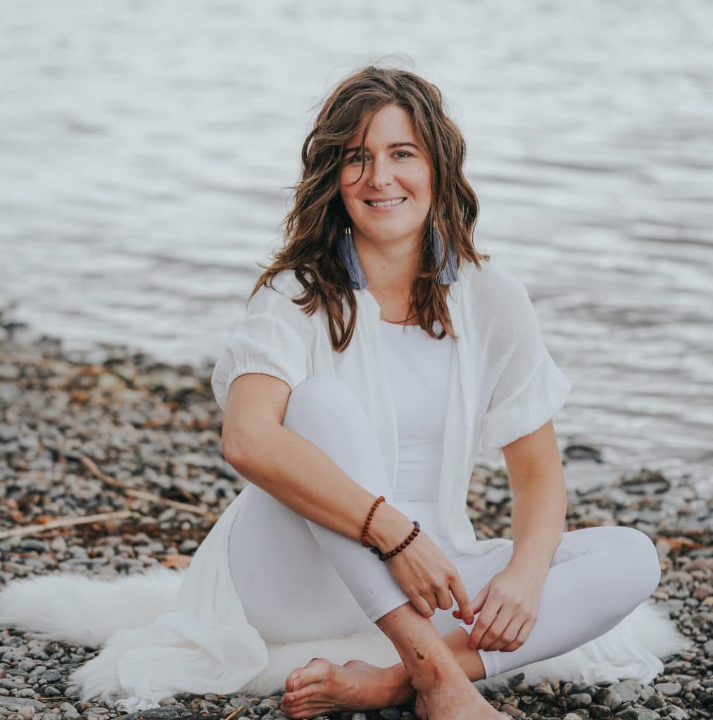 Erin Vivian sits on the edge of a river bank wearing all white. She is an energy healer certified in the emotion code and body code.