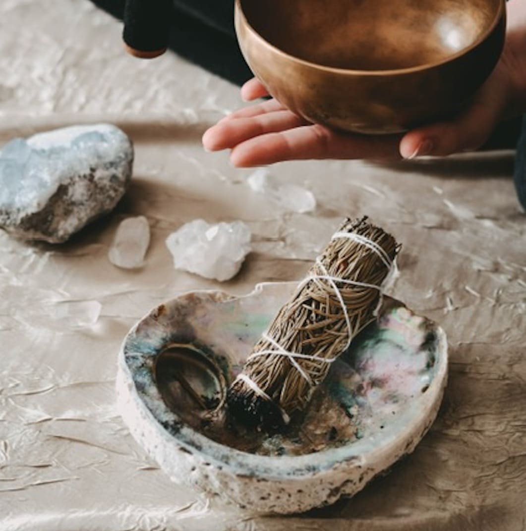 A shell bowl with a smudge stick burning inside it. A woman holds a singing bowl in her hand with crystals in the background.