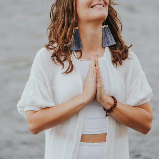 Erin Vivian standing on the river bank with her hands in prayer pose. She is smiling and looking up, and feeling grateful for her Energy Healing practice.